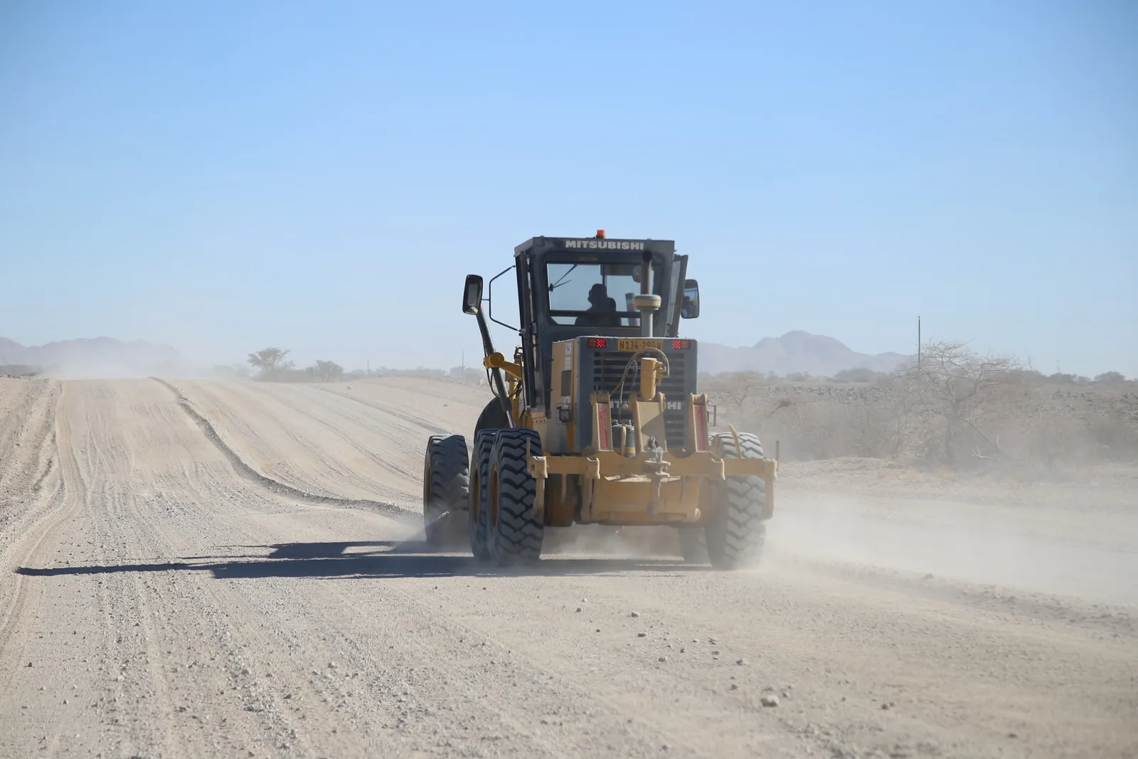 A yellow motor grader reshaping a gravel road in an arid landscape