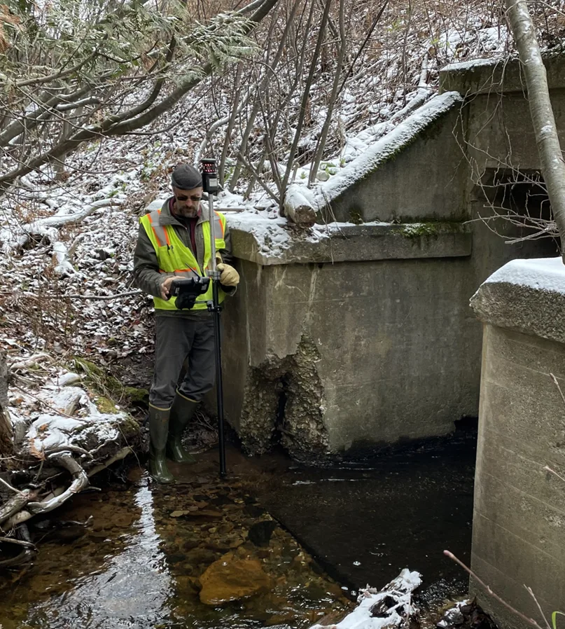 An engineer using survey equipment beside a concrete culvert in snowy woodland